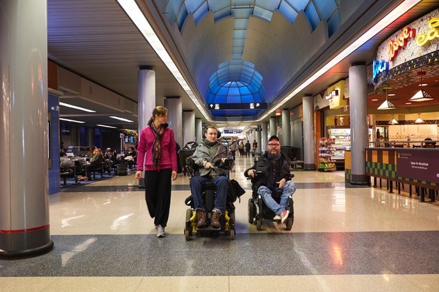 Two men in electronic wheelchairs and a female walking down the corridor of a busy airport.
