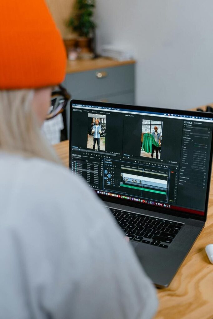 A young person with glasses and an orange beanie, focused on a laptop screen showing a video editing software interface with multiple clips and editing tracks visible.