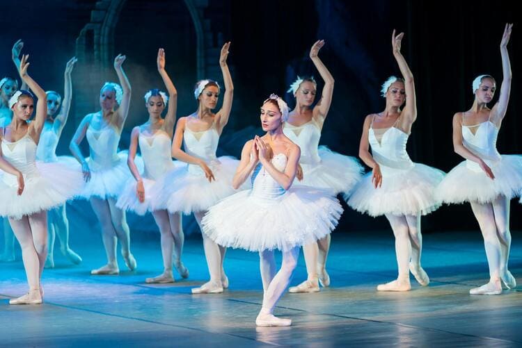 Group of ballet dancers on stage performing in white tutus, with a focus on one dancer in the foreground, against a softly lit blue background.