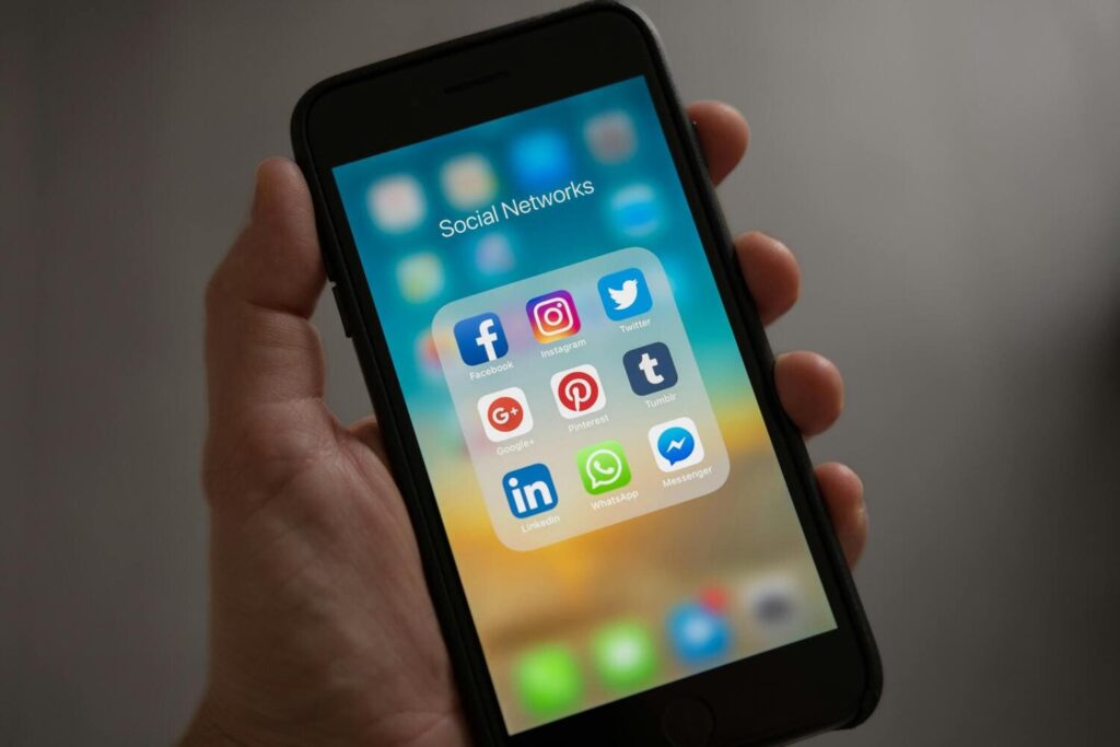 Close-up of a hand holding a smartphone displaying social media app icons including Facebook, Instagram, Twitter, Pinterest, LinkedIn, and WhatsApp on a colorful screen.